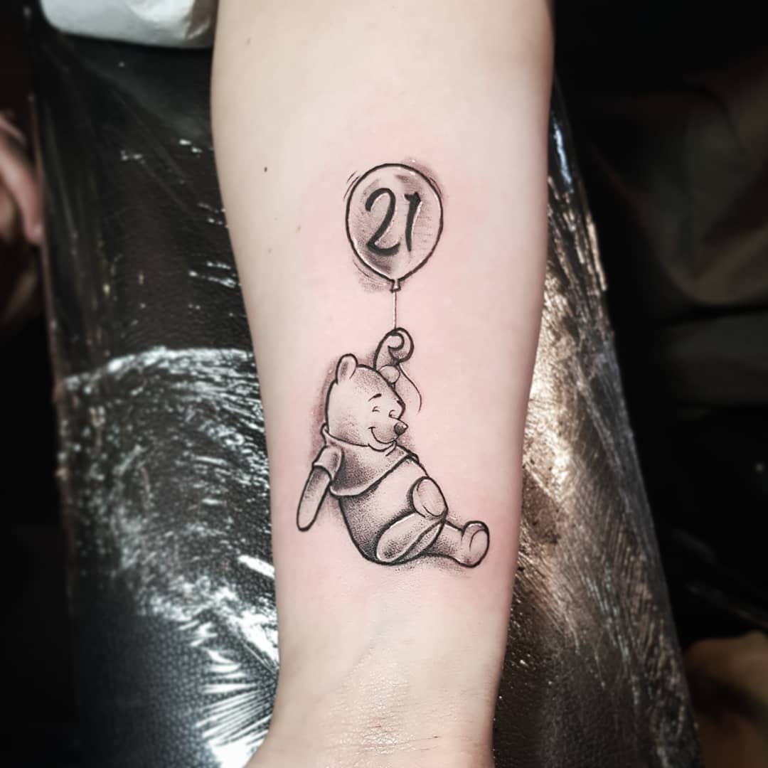 Tattoo of Winnie the Pooh floating with a balloon that has the number '21' on it, on a person's forearm